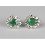 A pair of white gold, emerald and diamond earrings, each claw set with an oval cut emerald within