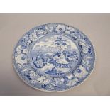 A blue and white plate printed with a pattern showing travellers resting beside a river and a drover