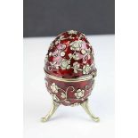 Russian style musical trinket box egg, raised on three feet, the red enamelled body set with paste
