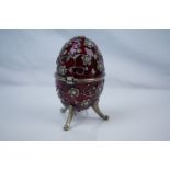 Russian style musical trinket box egg, raised on three feet, the red enamelled body set with paste