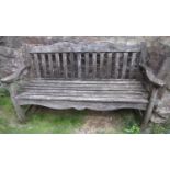 A well weathered teak three seat garden bench with shaped apron beneath a slatted seat and raised