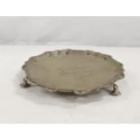 Silver scalloped edge waiter raised on three pad supports, with central inscription. 14cm.