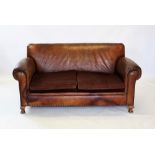 An early 20th century tan leather three piece lounge suite, the two seater settee with padded scroll