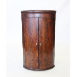 An early 19th century mahogany bow front hanging corner cupboard, the pair of flame mahogany doors