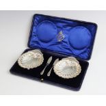 A cased pair of George V silver shell shaped butter dishes and knives by The Alexander Clark