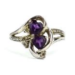 A white metal ring set two heart shaped purple coloured stones and small diamonds in a crossover