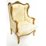 An early 20th century gilt wood framed wingback armchair, the frame with carved scrolling detail,