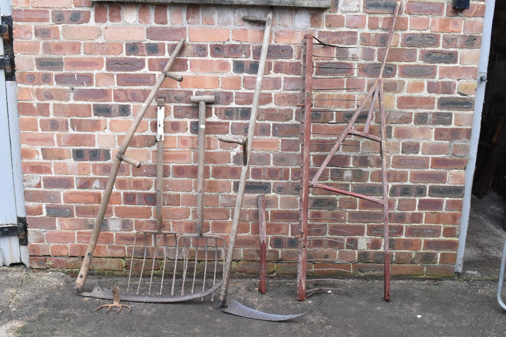 A vintage red painted wood and iron hay rake, two scythes, two potato forks, a fork and a fork head.