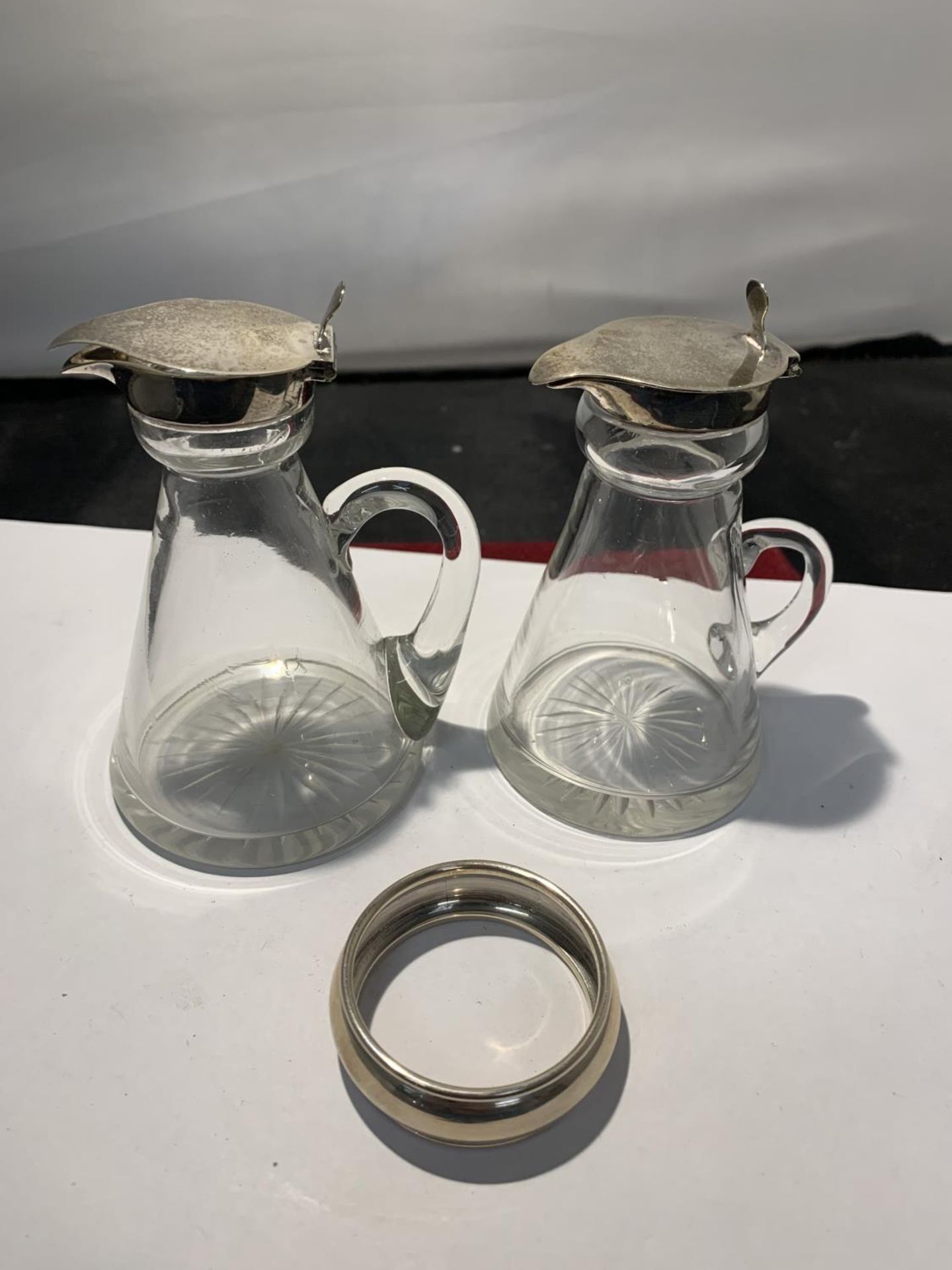 TWO GLASS JUGS WITH HALLMARKED BIRMINGHAM SILVER TOPS AND A SILVER NAPKIN RING