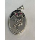 AN OVAL MARKED SILVER LOCKET WITH A RED STONE EYED OWL DESIGN