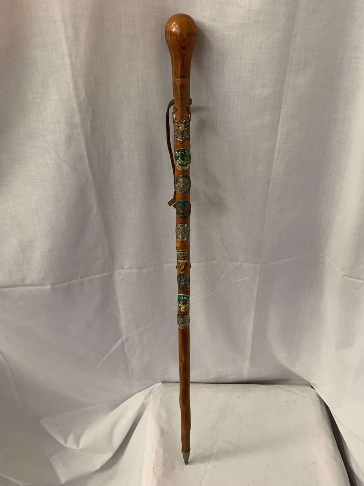 A WOODEN WALKING CANE WITH METAL TIP, LEATHER STRAP AND VARIOUS BADGES FROM THE ALPS