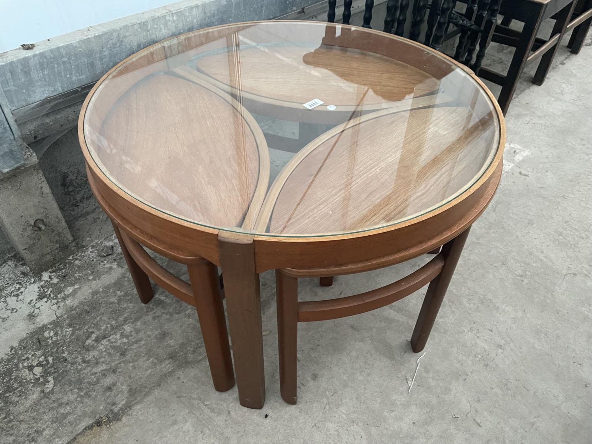 A RETRO TEAK CIRCULAR NEST OF FOUR TABLES WITH GLASS TOP AND THREE UNDER TABLES, OVAL IN SHAPE,