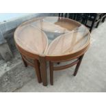 A RETRO TEAK CIRCULAR NEST OF FOUR TABLES WITH GLASS TOP AND THREE UNDER TABLES, OVAL IN SHAPE,