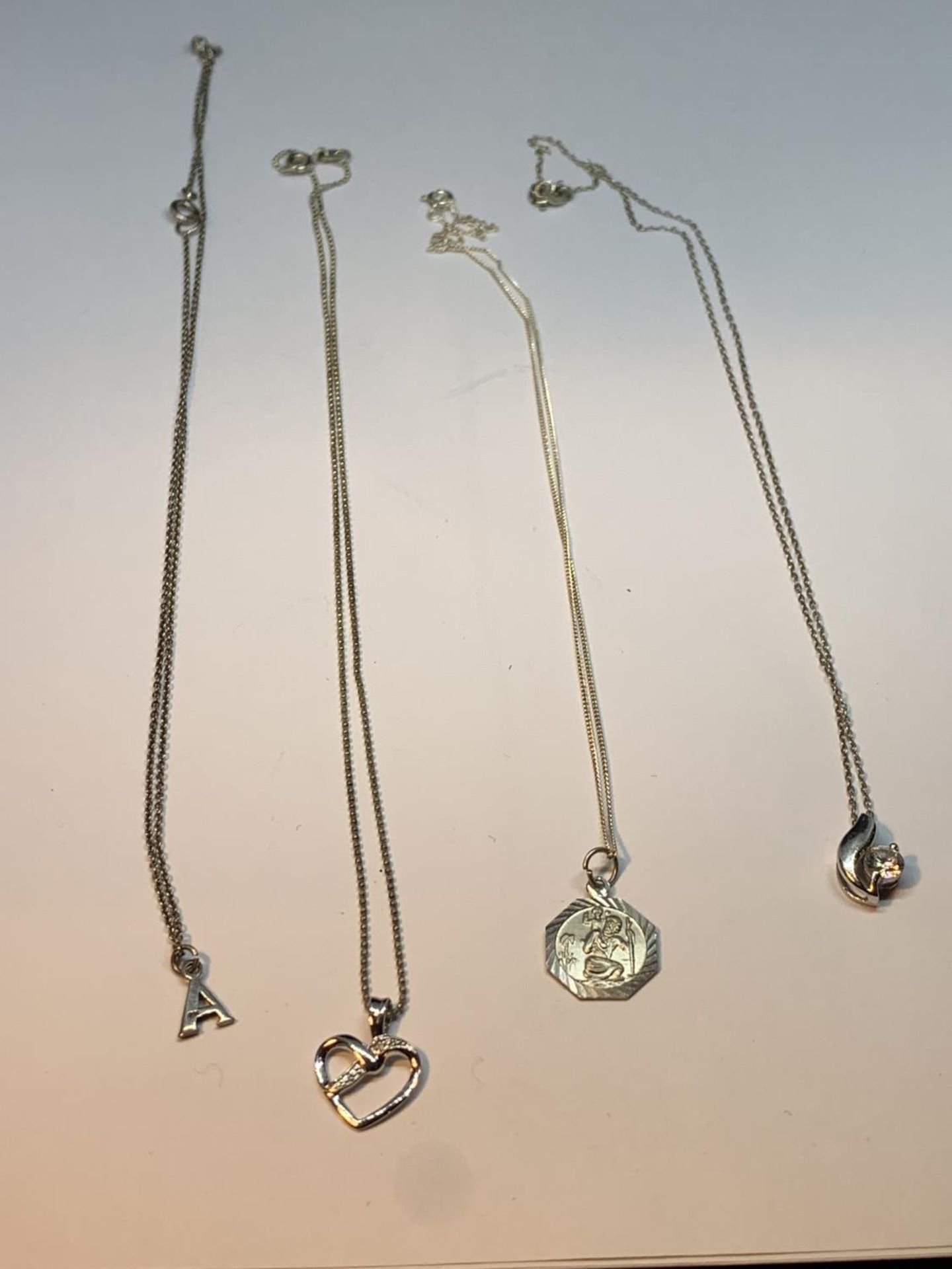FOUR SILVER NECKLACES WITH PENDANTS TO INCLUDE A CLEARSTONE FLOWER, HEART, ST CHRISTOPHER AND AN A