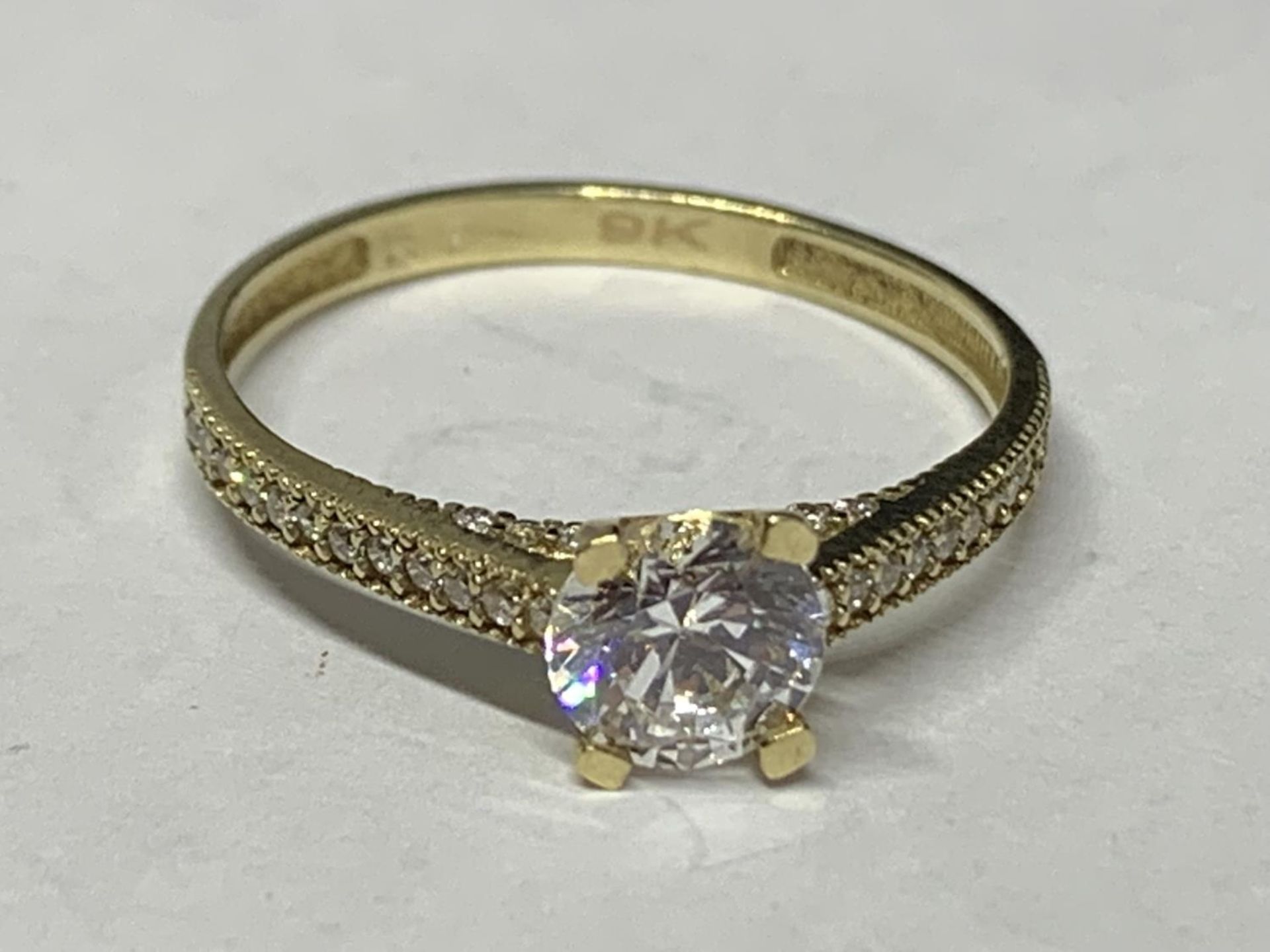 A 9 CARAT GOLD RING WITH A SINGLE CLEAR STONE AND CHIPS ON THE SHOULDERS SIZE O/P IN A