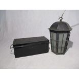 A metal hanging lantern light with glass panels, along with a black storage tin.