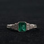 An 18ct white gold emerald and diamond art deco style ring. Central bezel set emerald with stepped