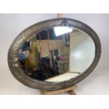 An oval arts and crafts brass framed mirror with hammered detail. Mirror has bevelled edge and