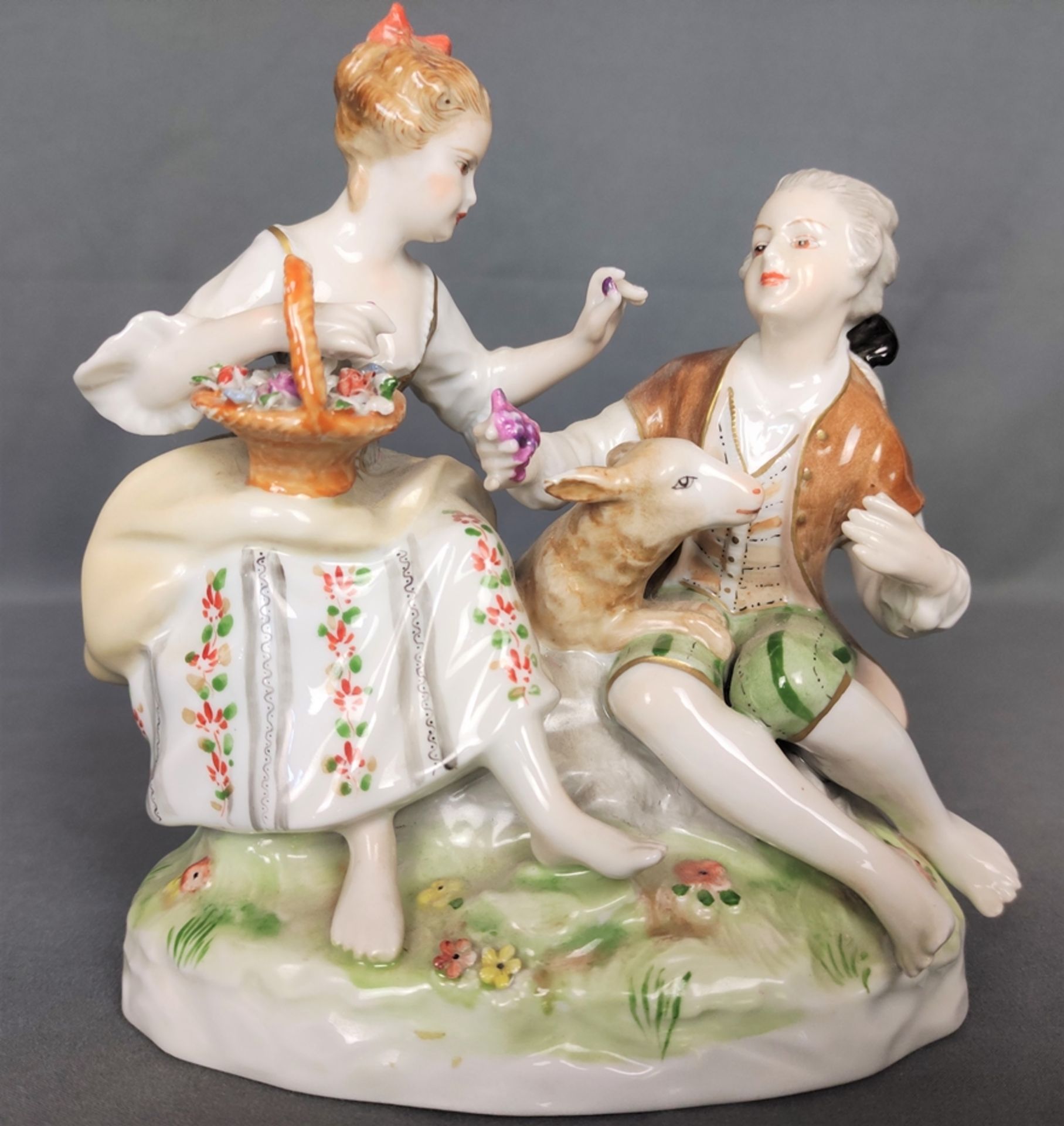 Porcelain group, couple sitting, between them a little sheep, her holding a flower basket, handing 