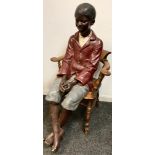 An early 20th century plaster life size model, of a black boy, seated, wearing a red jacket, blue