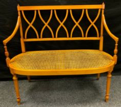 A Sheraton revival satinwood love seat, crossed splat back, open arms, turned fore legs, caned seat,