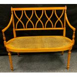 A Sheraton revival satinwood love seat, crossed splat back, open arms, turned fore legs, caned seat,