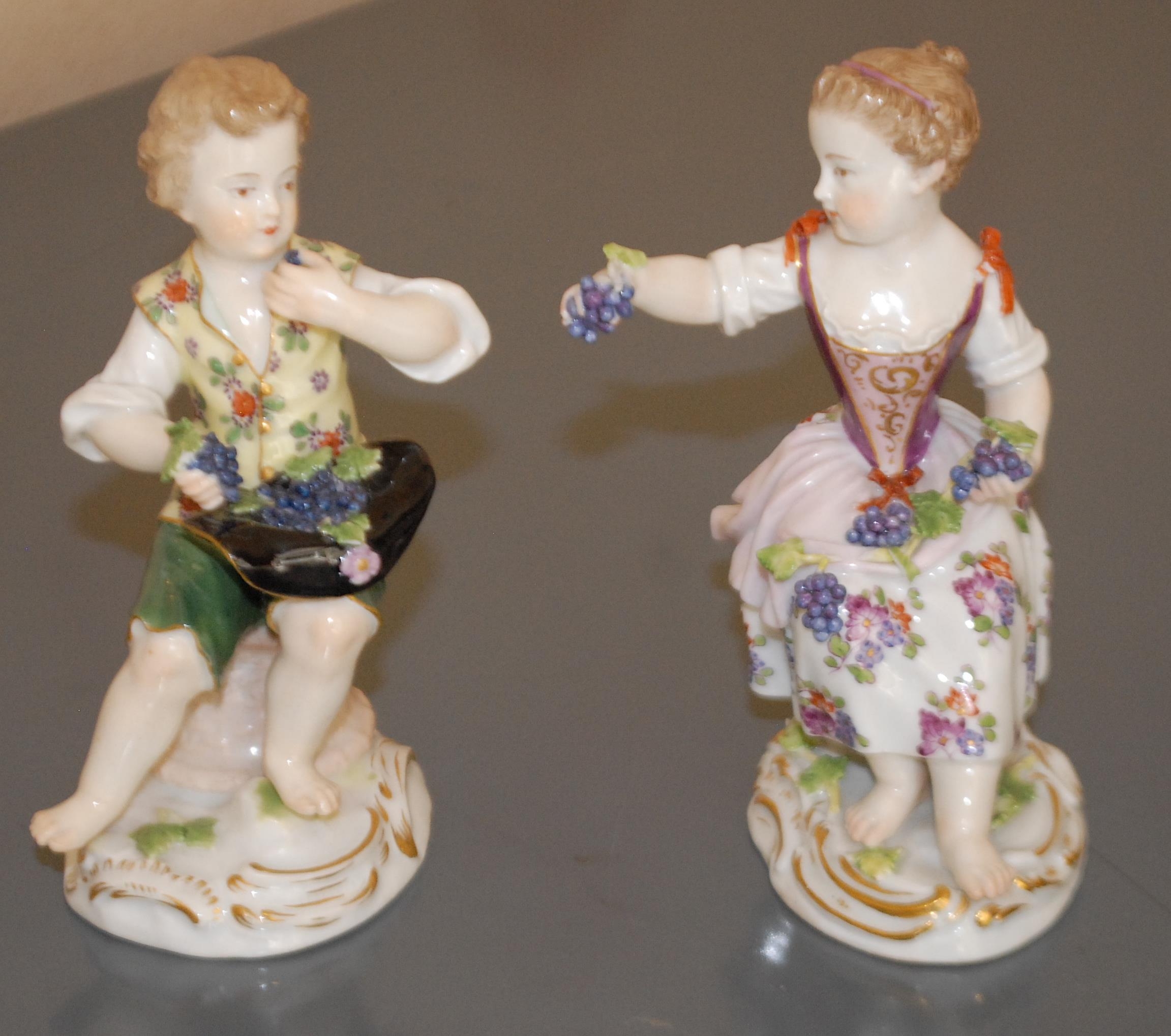 A pair of Meissen figures, of a girl and a boy, both bare foot, seated, she with grapes in her