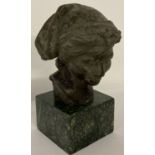 A small bronze bust of a boy wearing a hat, mounted on a square shaped green marble base.