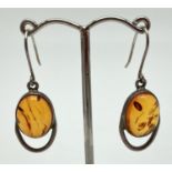 A pair of modern design drop style earrings set with an oval cabochon of amber. Silver marks to