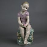 Porzellanfigur - Liebreizende Mädchenfigur, auf einem Steinquader sitzend, mit Rosenblüten in den H