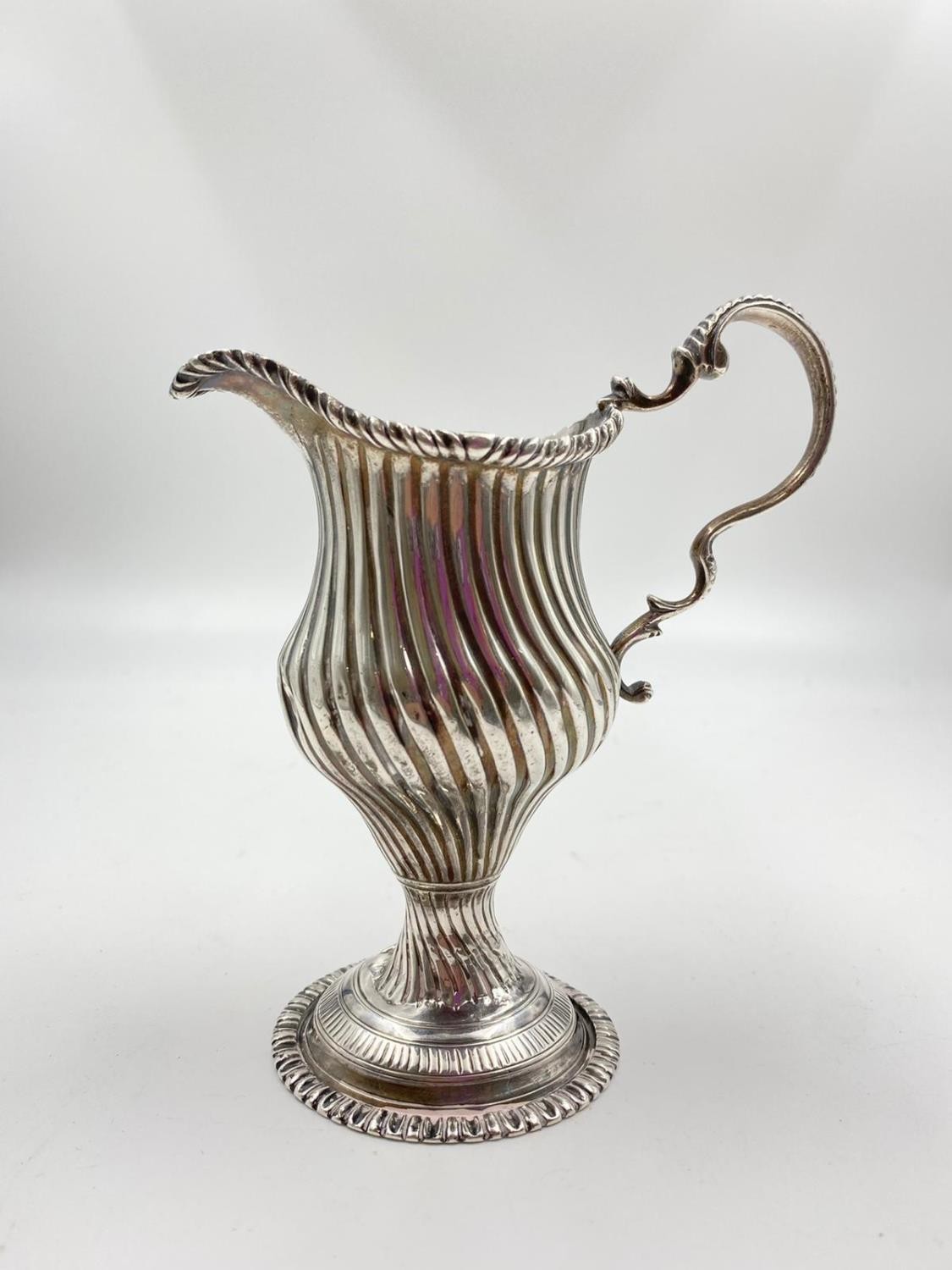 A finely detailed silver creamer in ribbed swirl pattern with curved handle on a pedestal base. 14cm
