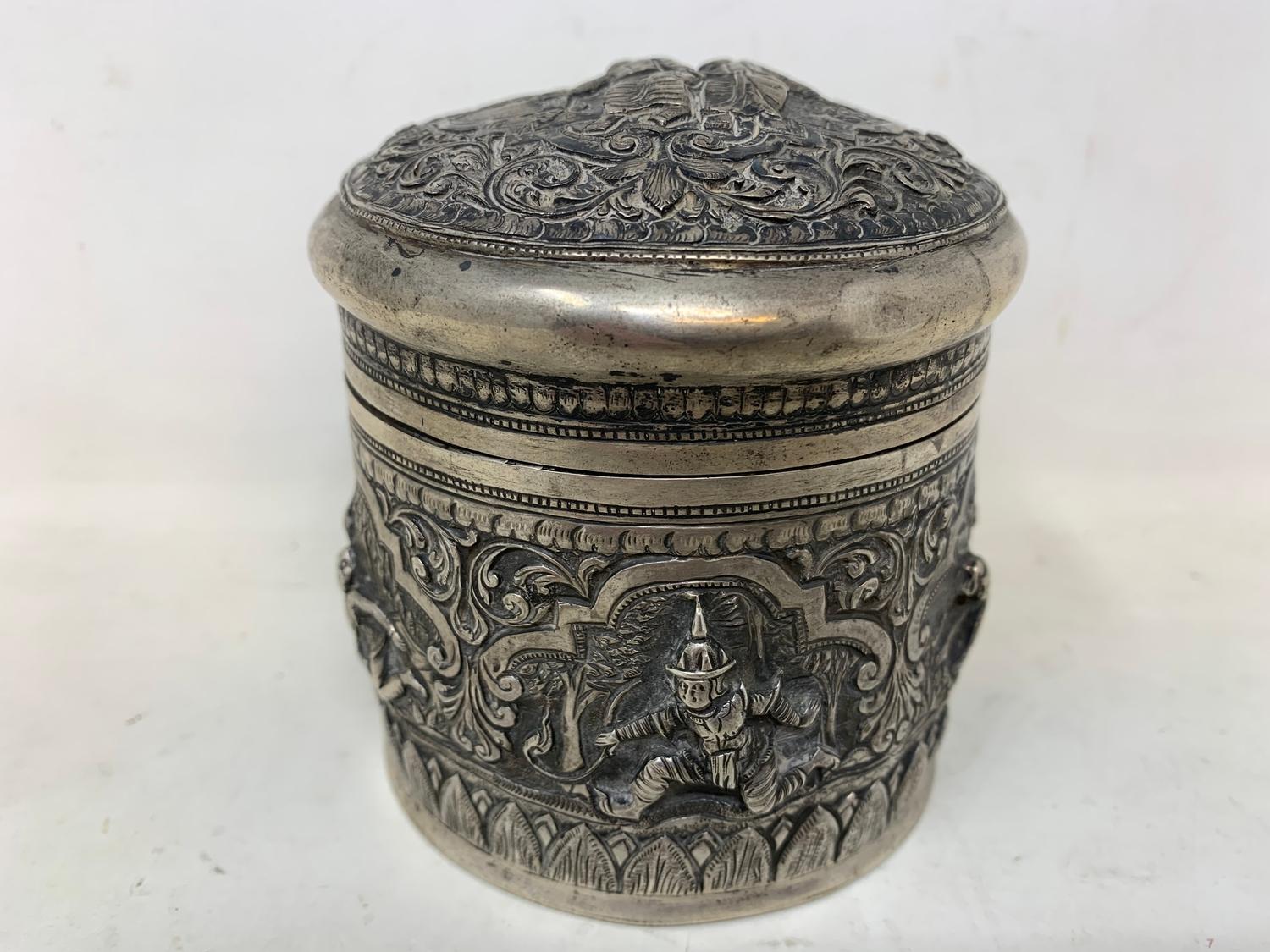 A Burmese silver coloured metal cylindrical box and cover, embossed figures, the base engraved a