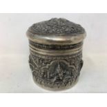 A Burmese silver coloured metal cylindrical box and cover, embossed figures, the base engraved a
