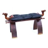 A camel saddle stool with black leather studded cushion on a brass studded and carved wood frame