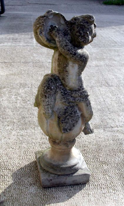 A well weathered reconstituted stone figure of a cherub playing a tambourine, seated on a column,