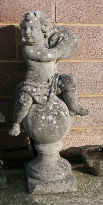 A well weathered reconstituted stone figure of a cherub playing a pair of cymbals, seated on a