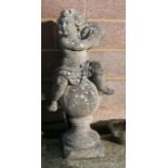A well weathered reconstituted stone figure of a cherub playing a pair of cymbals, seated on a