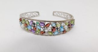 A multi gemstone and silver scrolling design pierced bangle. Set with forty eight mixed cut