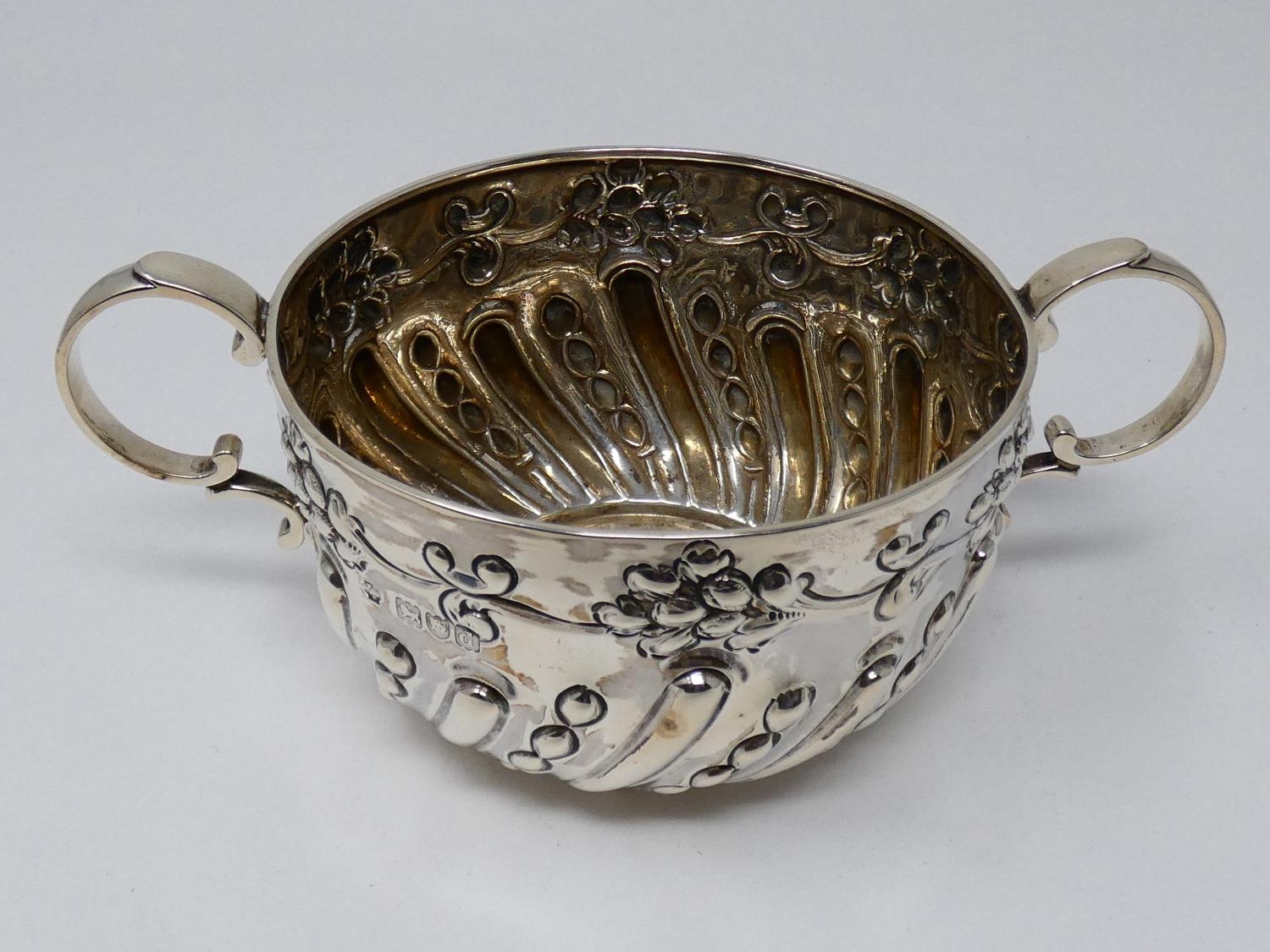 A two handled Victorian repousse design silver porriger with floral design and scrolling handles.