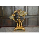 A gilt metal three branch table lamp in the form of a cherub supporting scrolling foliate arms