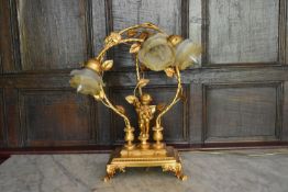 A gilt metal three branch table lamp in the form of a cherub supporting scrolling foliate arms