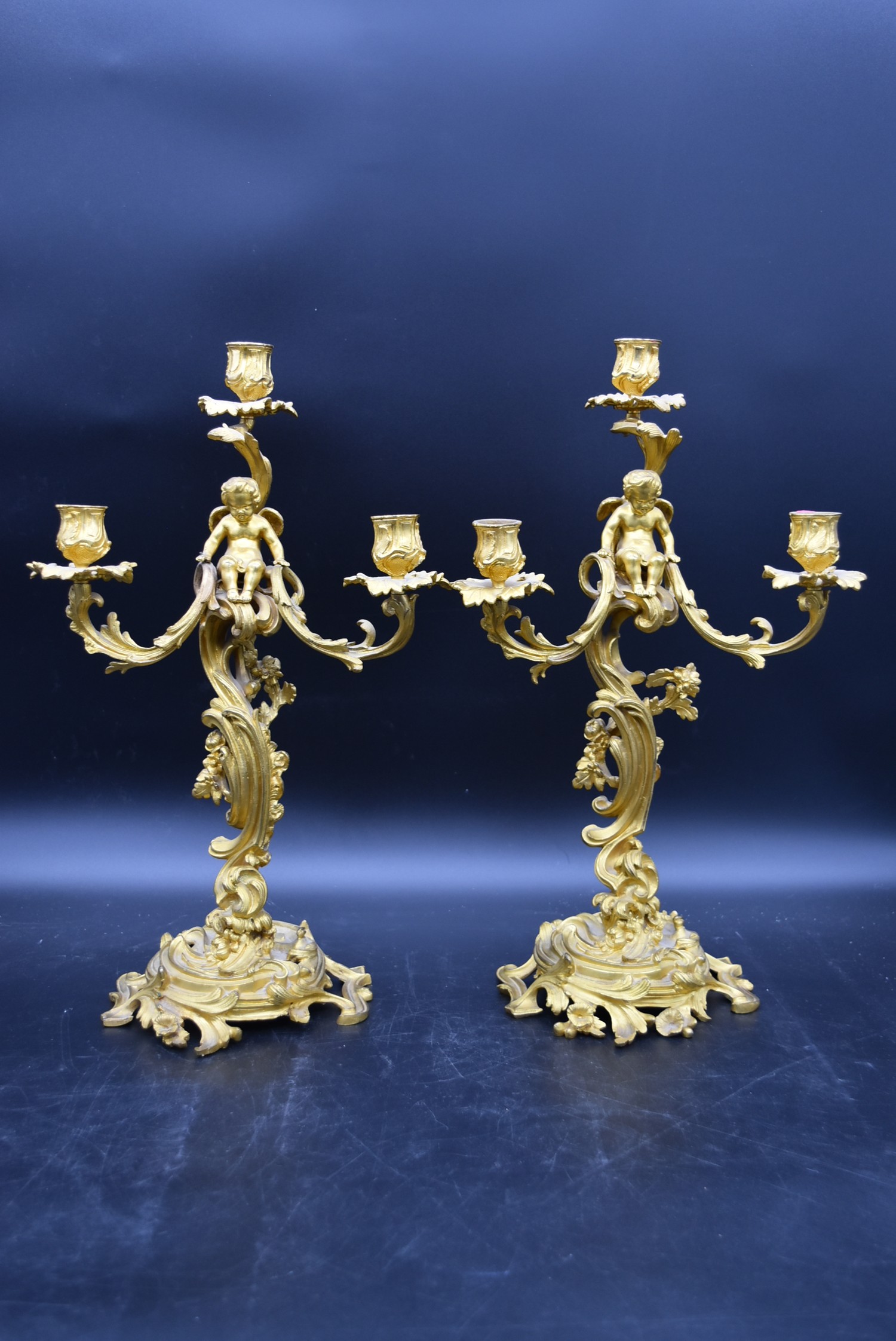 A pair of gilt metal three branch table candelabras with cherub surmounts raised on Rococo scrolling