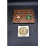 An embossed copper armorial plaque and a glazed tile depicting medieval wine making. H.25 W.33cm