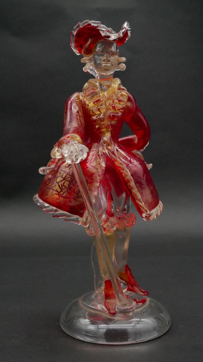 A vintage Murano gold flecked red glass figure of a Venetian gentleman with cane, on a clear round