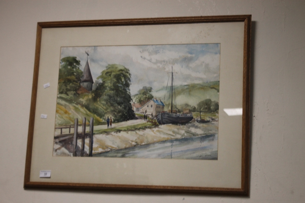 A FRAMED AND GLAZED WATERCOLOUR DEPICTING BOATS ON THE EDGE OF A VILLAGE SIGNED TO THE LOWER