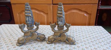 A pair of brass fire dogs depicting a sailing ship above two oversized fish.