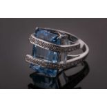 A white metal, diamond, and blue topaz ring, set with a rectangular-cut topaz within a curved double