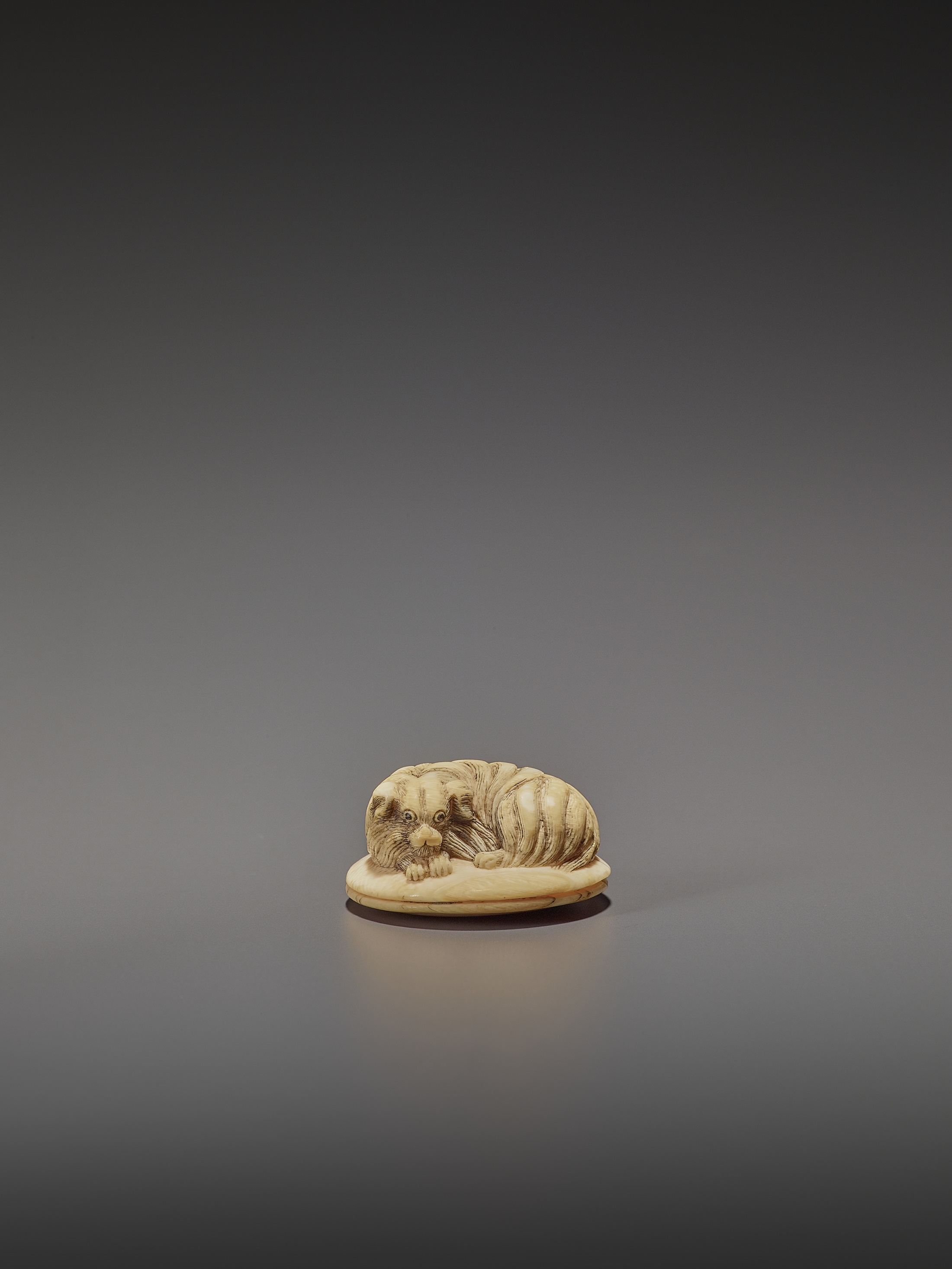 AN IVORY NETSUKE OF A SHAGGY DOG ON A ZABUTON