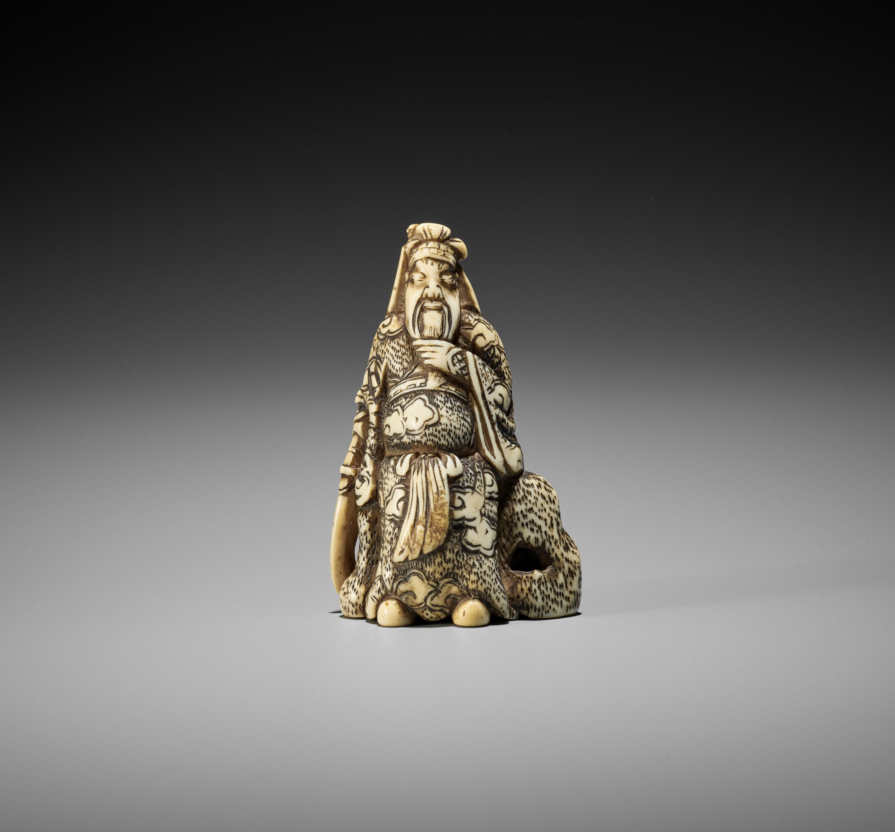 AN IVORY NETSUKE OF KAN'U LEANING AGAINST A ROCK