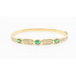 A diamond and emerald 14ct gold hinged bracelet, comprising three oval emeralds rub over set within